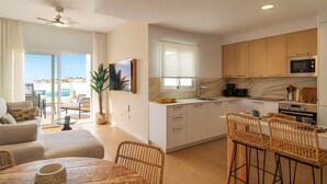 A modern kitchen with white cabinets, a built-in oven, and a microwave. There is a dining area with wicker chairs and a table.