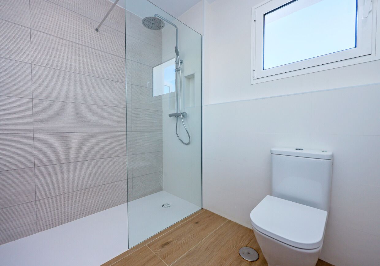A modern bathroom with a glass shower enclosure, a white toilet, and a window above the toilet.