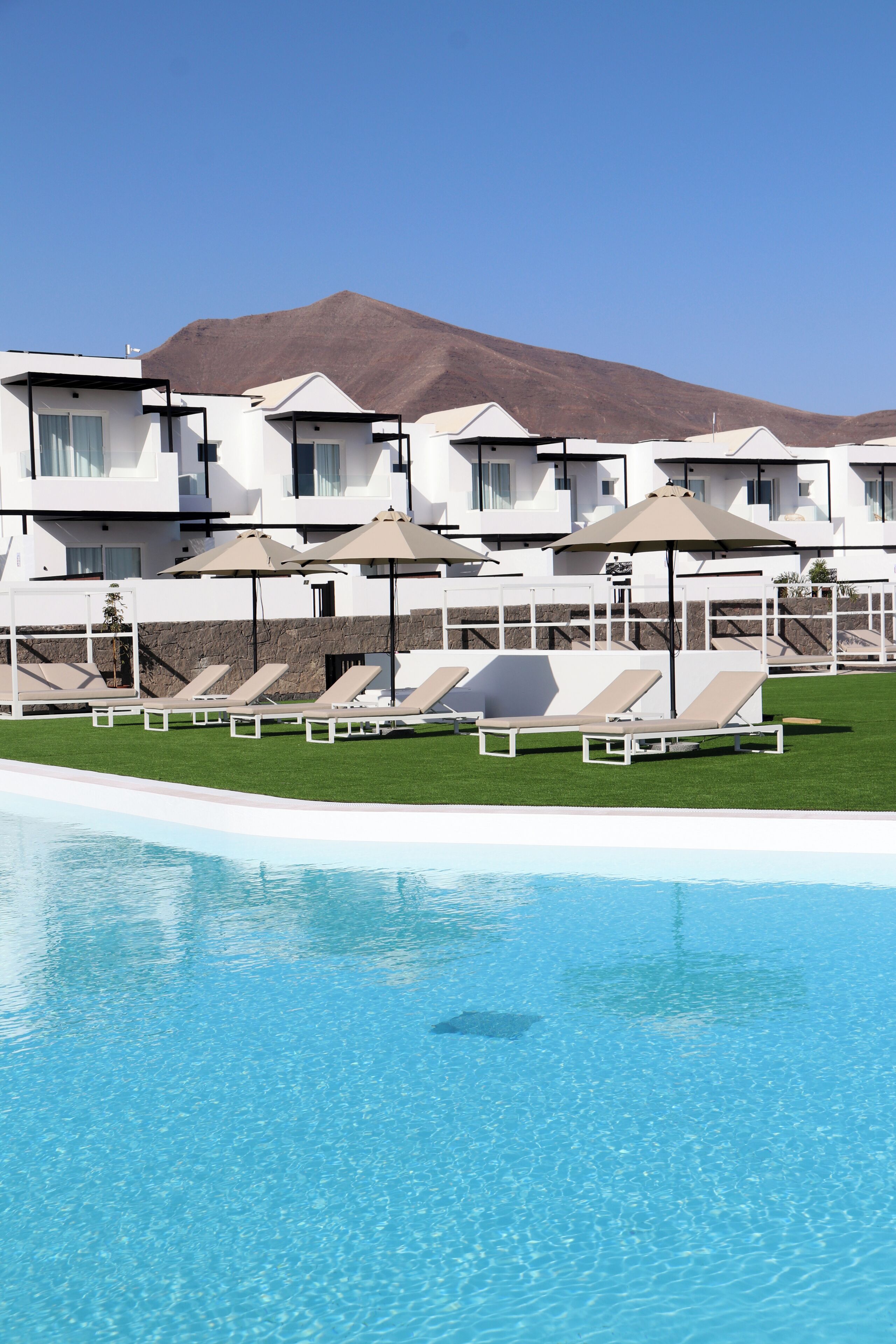 Zona de piscina con sillas de descanso y sombrillas, una piscina azul cristalina y una montaña al fondo.