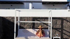 A modern outdoor bed with a person sitting on it, a stone wall, and a white building in the background.