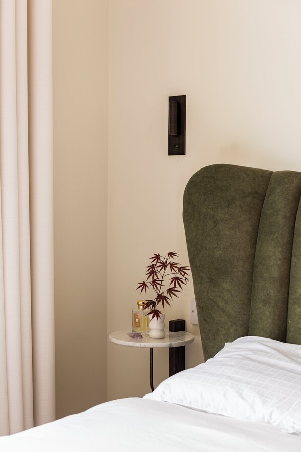 A hotel room with a bed, a small round table with a plant and a bottle, a wall-mounted light, and a green upholstered headboard. - Warm, Comfortable and beautifully decorated! (Dublin)