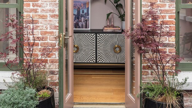 A brick building with a wooden door, two potted plants, and a cityscape poster.