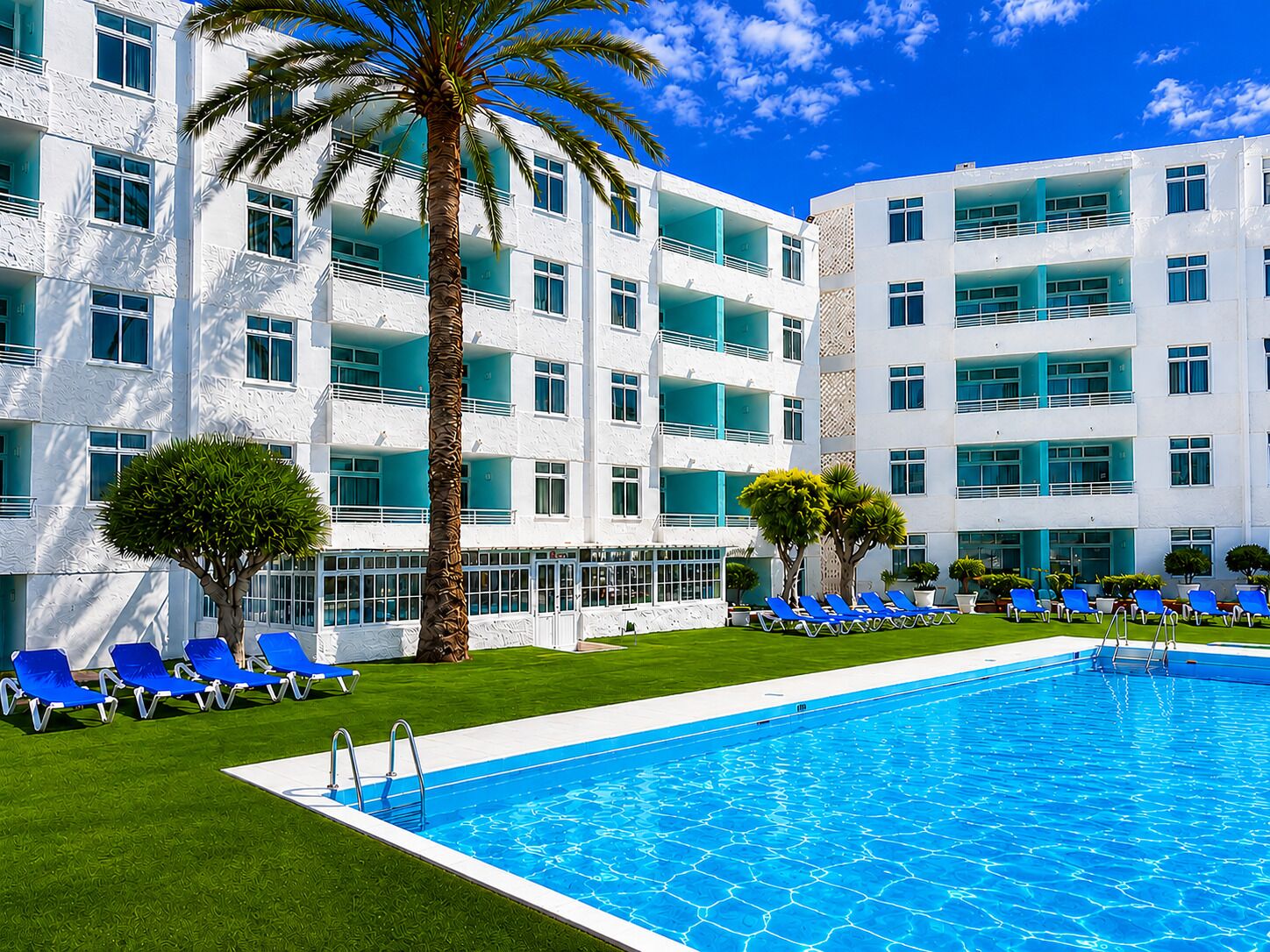 A hotel with a swimming pool, blue lounge chairs, and palm trees.
