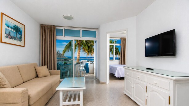 Ein Hotelzimmer mit einem beigen Sofa, einem Glastisch, einem weißen Schrank, einem Flachbildfernseher und Blick aufs Meer.