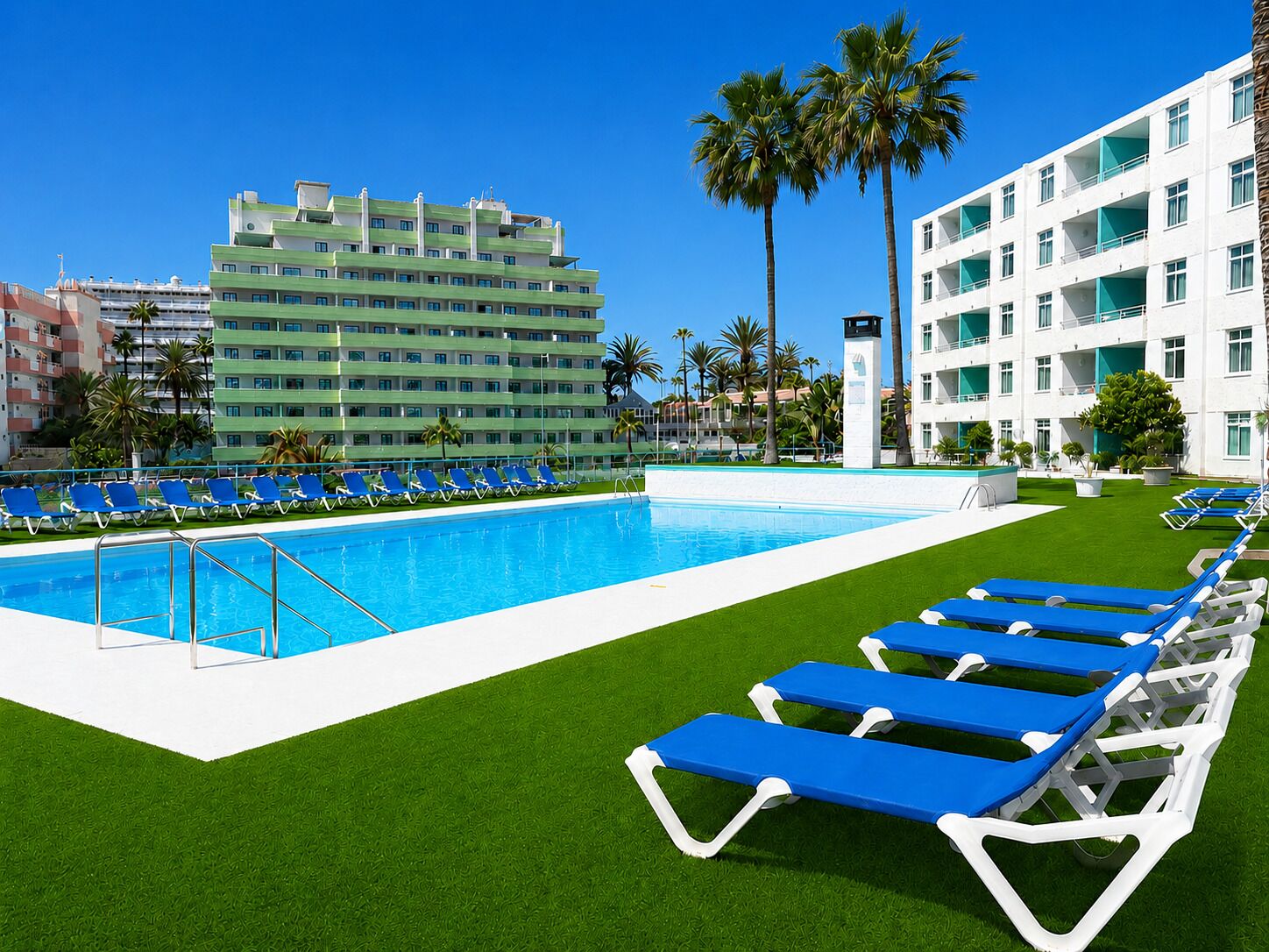 Una piscina con sillas reclinables azules, un césped verde y un edificio de varios pisos al fondo.
