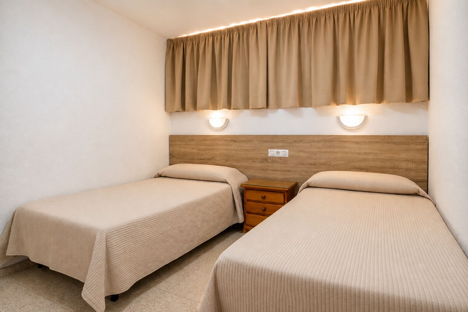 Quarto duplo com duas camas, cabeceira de madeira, mesa de cabeceira e luminárias fixadas na parede.