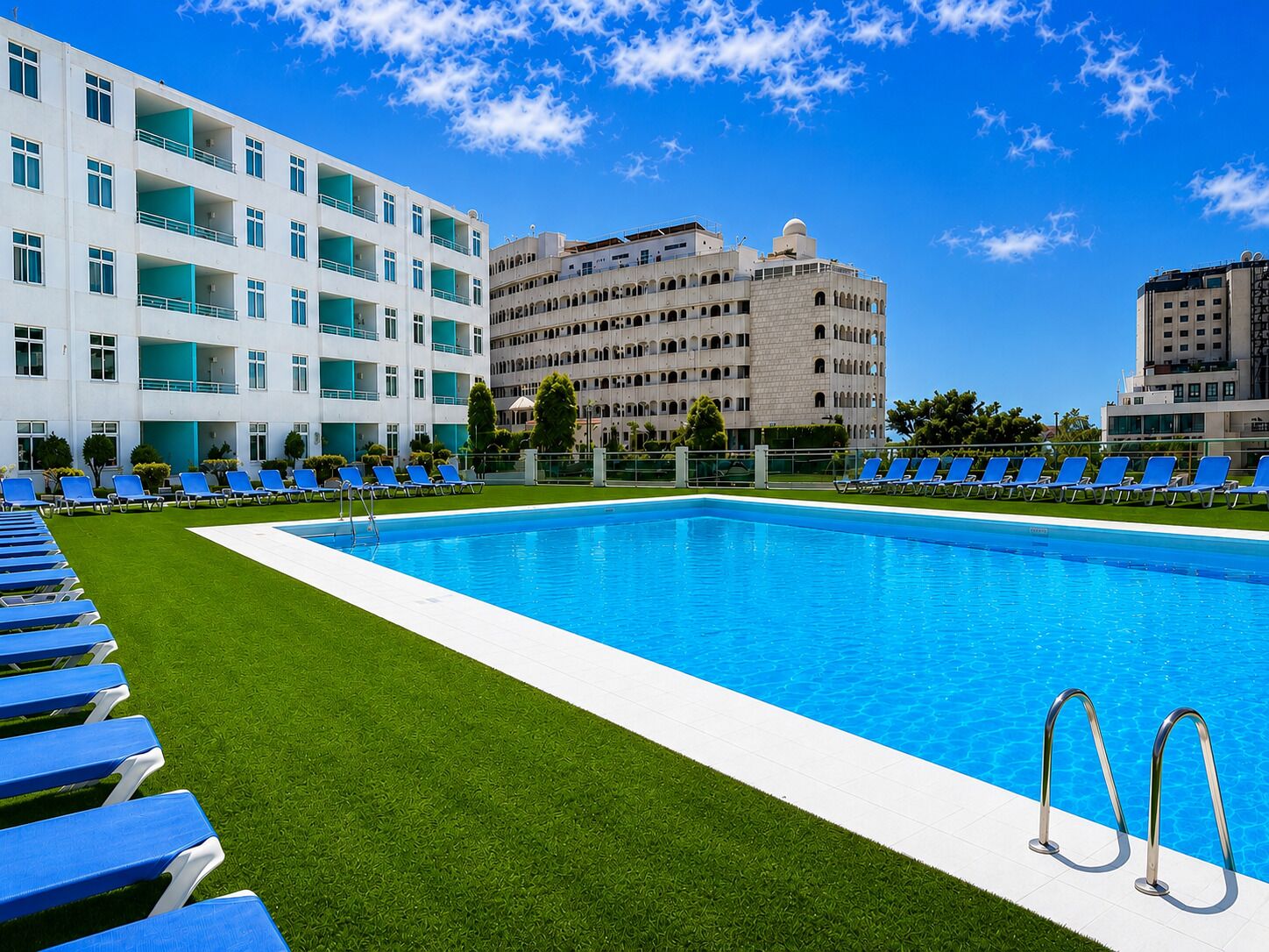 Una piscina con sillas reclinables azules, un área verde y un edificio con varios balcones y una cúpula.