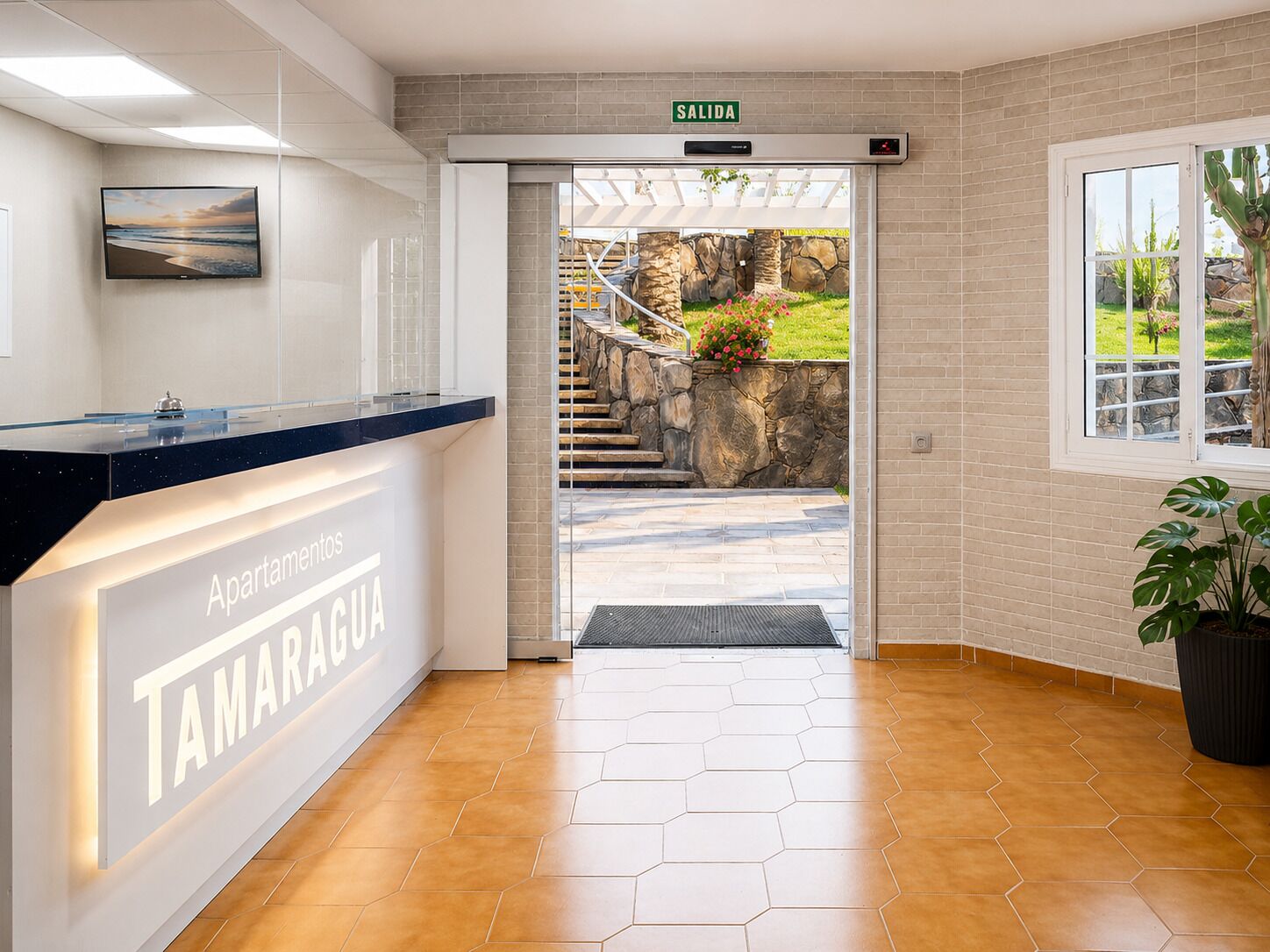 Un espace d’accueil avec une enseigne indiquant « Apartamentos TAMARAGUA », un comptoir de réception, un escalier menant à l’extérieur et une plante en pot.
