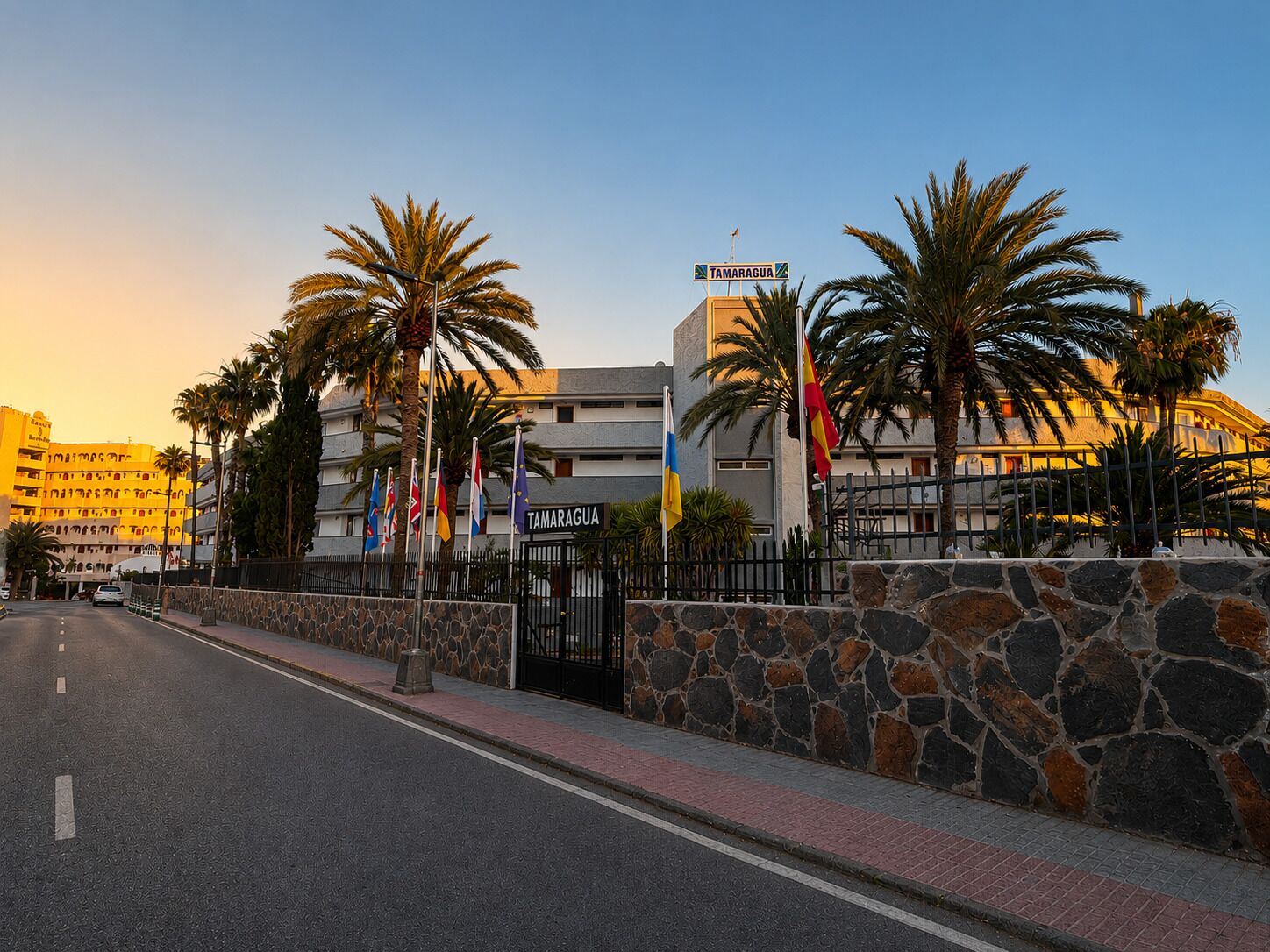 A hotel with a stone wall, palm trees, and multiple flags.