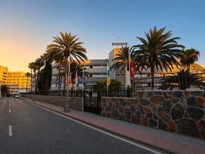 A hotel with a stone wall, palm trees, and multiple flags. - APARTAMENTOS TAMARAGUA (San Bartolome de Tirajana)