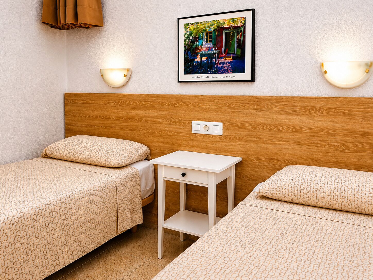 Ett hotellrum med två sängar, ett litet sängbord, en vägglampa och en inramad tavla på väggen.