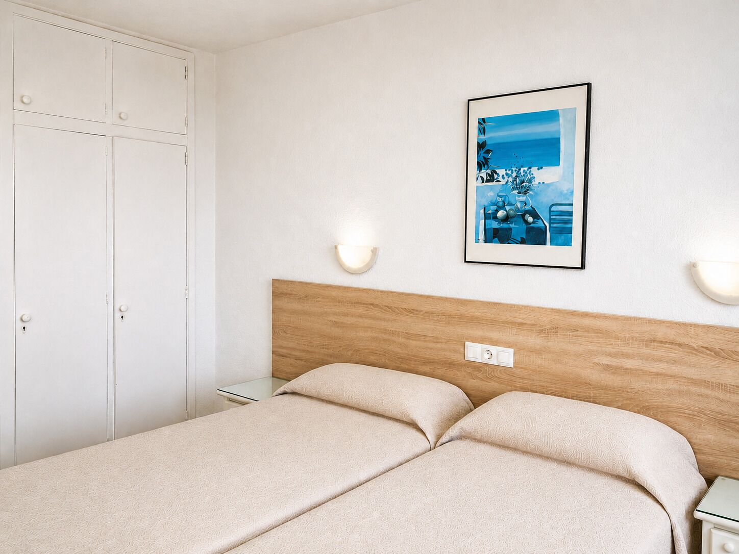 Un dormitorio con cabecera de madera, cama con ropa de cama color beige, armario blanco y un cuadro en la pared.