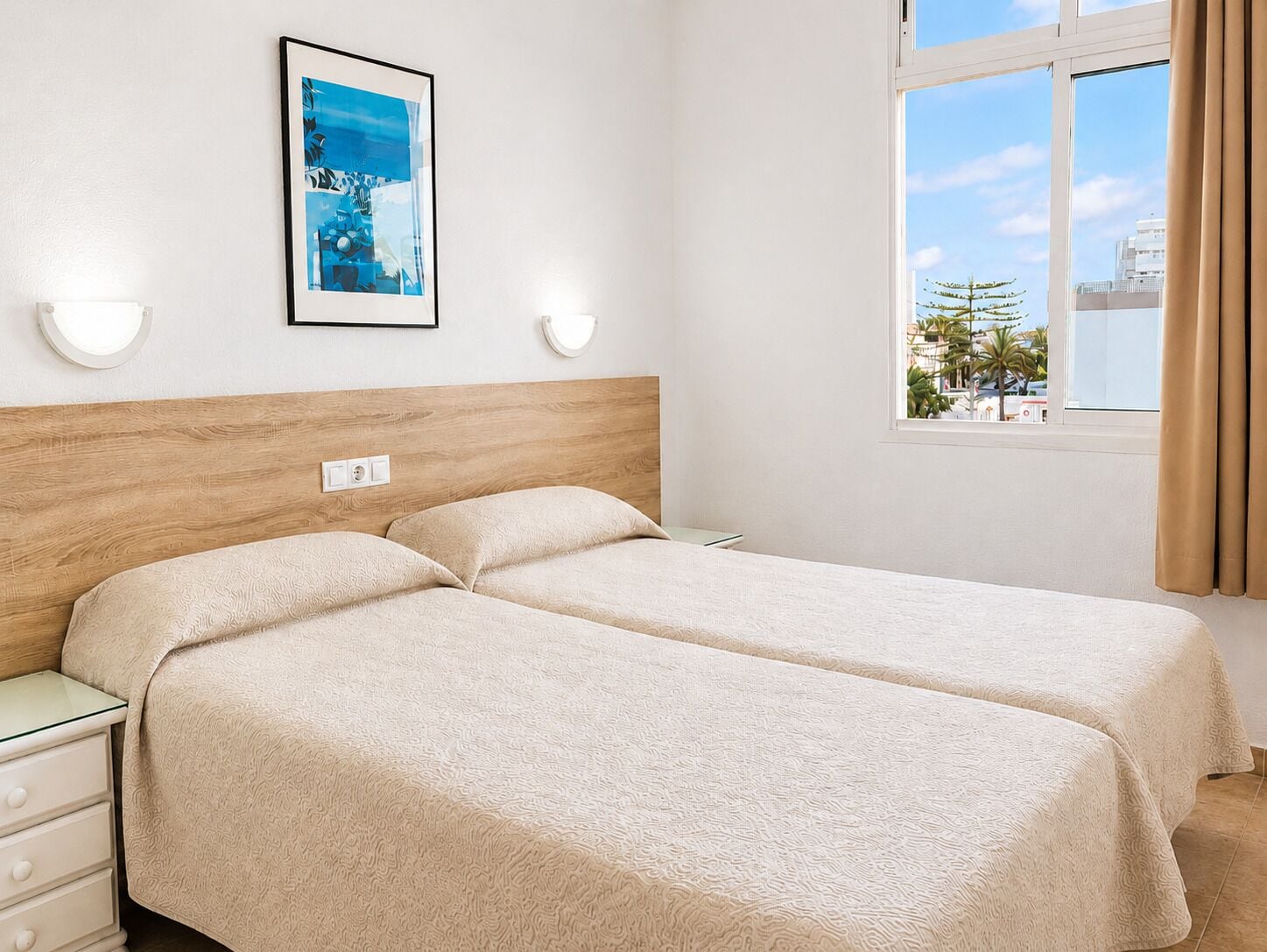 A hotel room with two beds, a wooden headboard, a window with a view of palm trees, and a framed picture on the wall.