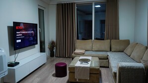 Un salon moderne avec un téléviseur à écran plat diffusant une publicité Netflix, un canapé, une table basse en verre et une grande fenêtre avec des rideaux.