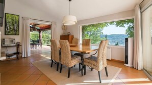 A dining area with a large table and wicker chairs, a pendant light, and a view of greenery and hills. - Country house with private pool, quiet comfort & nature (Seillans)