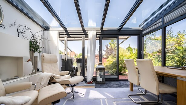 Een moderne kamer met glazen dak, een bureau, twee stoelen, een potplant en uitzicht op groen buiten.