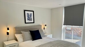 A modern bedroom with a bed, bedside tables, a nightstand, a framed picture on the wall, and a window with a blind. - By Allerton Manor & Beatles Spots | Free Parking (Liverpool)