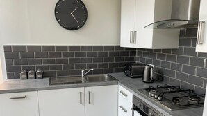 A modern kitchen with white cabinets, a black tiled backsplash, a stainless steel sink, a gas stove, and a black wall clock. - By Allerton Manor & Beatles Spots | Free Parking (Liverpool)
