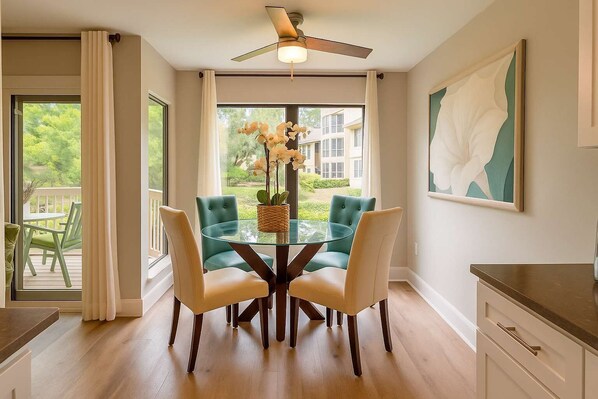A dining area with a glass table and four chairs, a ceiling fan, and a view of a green landscape through large windows.