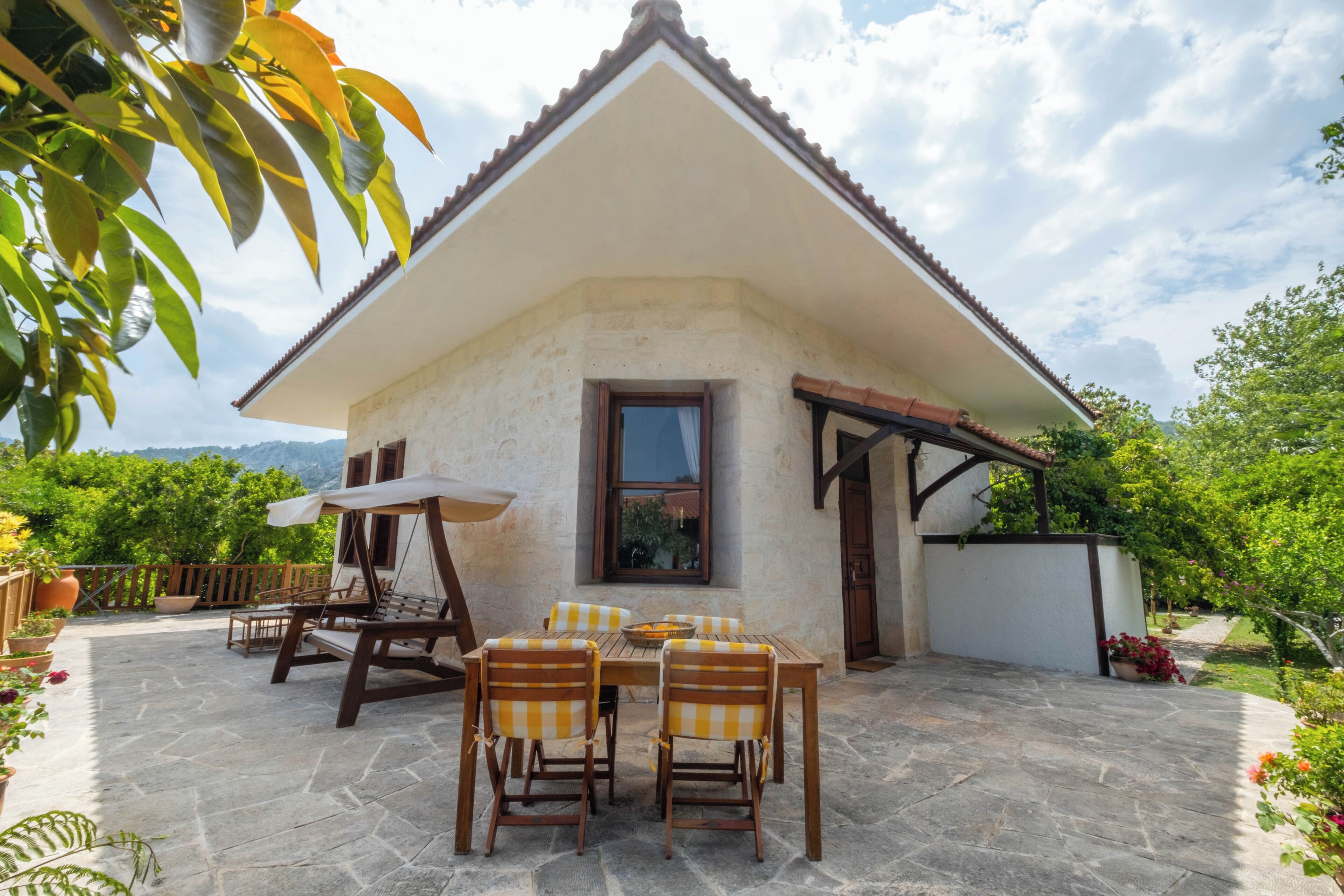 Une petite maison en pierre avec un toit en tuiles, une terrasse avec une table et des chaises, et un jardin agrémenté de plantes en pot.