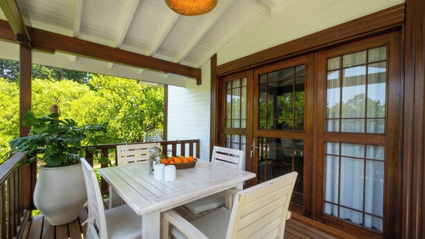 Une terrasse extérieure couverte, avec une table en bois et des chaises blanches, entourée de grandes fenêtres et agrémentée d’une plante en pot.