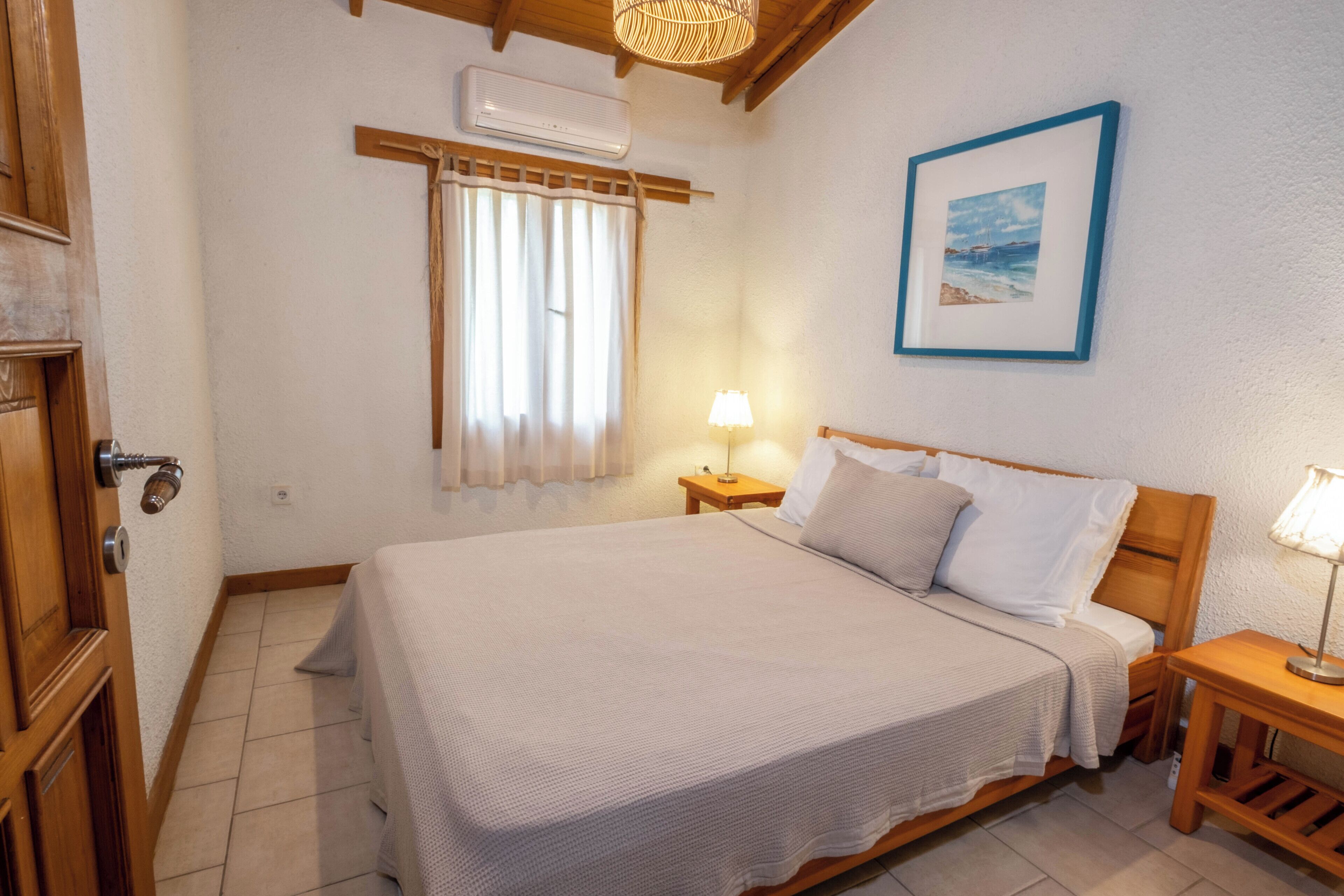 Quarto com cama, mesas de cabeceira, janela com cortinas, ar condicionado fixado na parede e um quadro emoldurado.