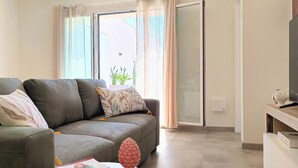 A living room with a grey sofa, a small table with a book and a decorative item, a TV on a stand, and a sliding glass door leading outside.