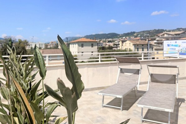A rooftop terrace with lounge chairs, a view of a town, and a plant in the foreground.