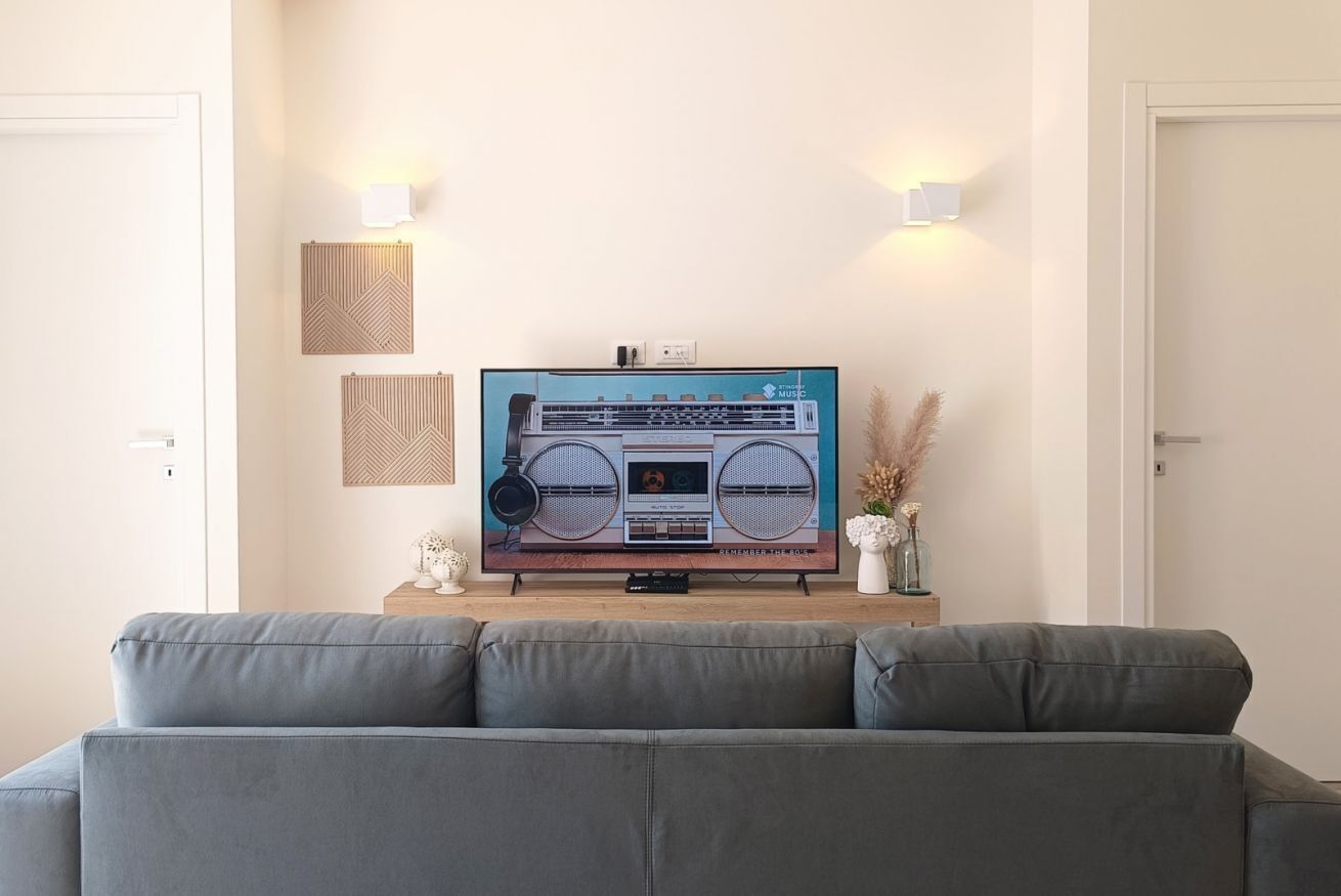 Een woonkamer met een grote flatscreen televisie op een houten standaard, een grijze bank en twee wandlampen.