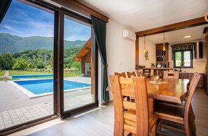 Uma sala de jantar com mesa e cadeiras de madeira, uma cozinha com eletrodomésticos modernos e vista para uma piscina e montanhas.