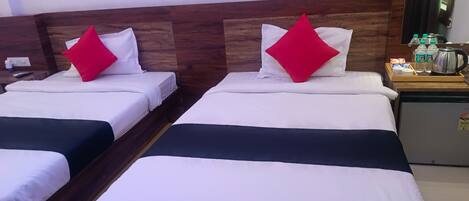 Habitación de hotel con dos camas, cada una con ropa blanca y almohadas rojas. Se observa un cabecero de madera, una mesita con artículos de tocador y un refrigerador.