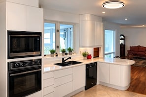 A modern kitchen with a built-in oven, microwave, and dishwasher. There are potted plants on the windowsill and a red vase on the countertop.
