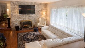 A living room with a stone wall, a flat-screen TV, a fireplace, a white sofa, and a large window with sheer curtains. - L`urbaine. Piscine creusée. (Trois-Rivières)