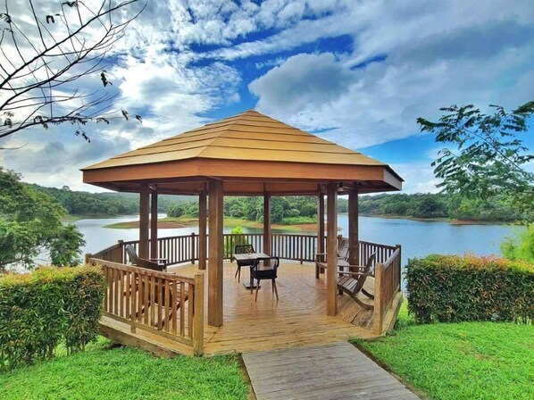 A wooden gazebo with a roof, surrounded by a wooden deck and a railing, overlooking a lake and greenery.