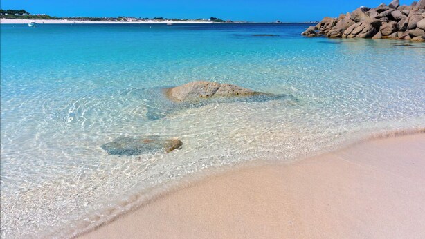 Une plage avec une eau turquoise et limpide, un rivage sablonneux et de gros rochers dans l’eau.