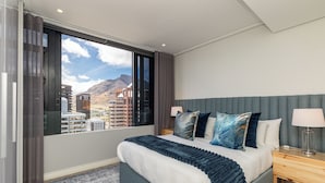 Une chambre d’hôtel moderne avec un grand lit, une table de chevet et une vue sur les immeubles et les montagnes, visible à travers une grande fenêtre.