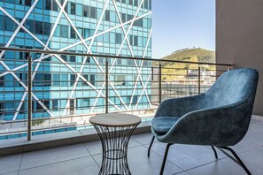 A balcony with a modern chair and a small round table, overlooking a building with a geometric glass facade and a view of a hill.