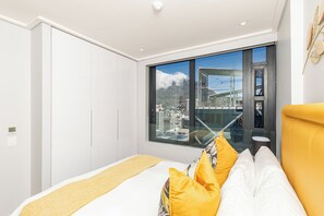 Una habitación de hotel moderna con un ventanal grande, una cama con ropa de cama amarilla y blanca, y vistas a la ciudad.