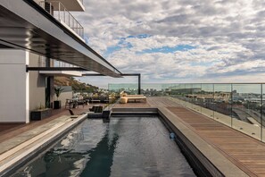 Una piscina en la azotea con vista a la ciudad, una terraza de madera y barandilla de cristal.