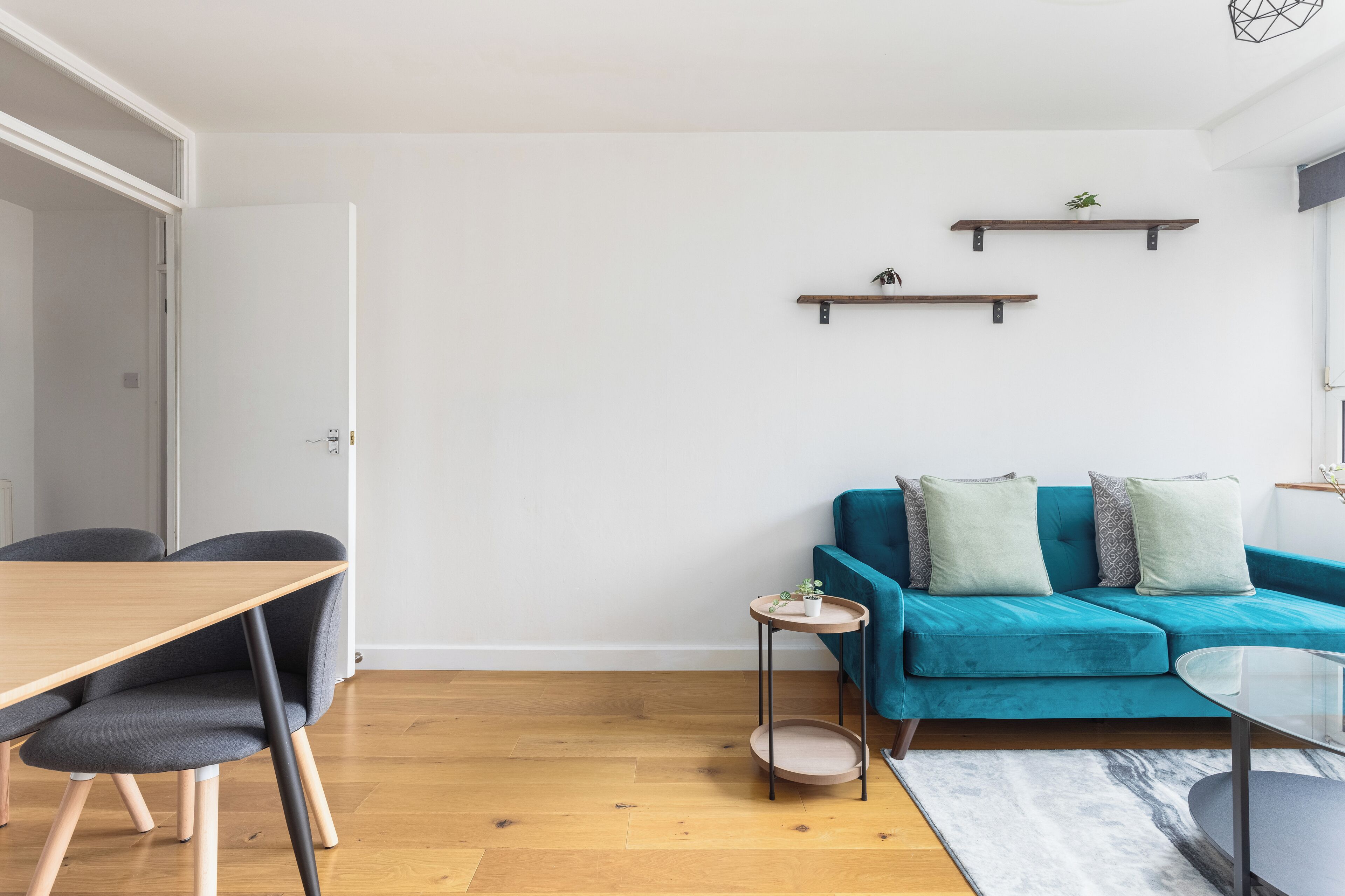 Un salon moderne avec une table à manger en bois, des chaises gris foncé, un canapé turquoise agrémenté de coussins et deux étagères murales ornées de plantes.