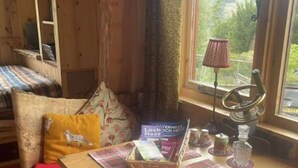 Ein gemütliches Blockhaus-Interieur mit einem Holztisch, einer Bank, einem Fenster mit Aussicht und einem kleinen Regal mit Büchern und einer Lampe.