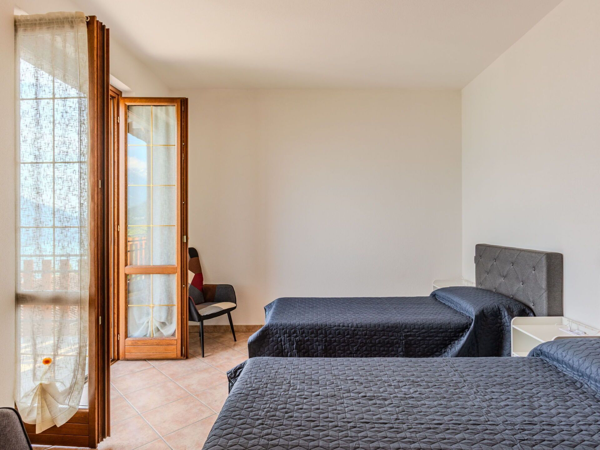 Quarto com duas camas, uma cadeira e porta de vidro deslizante para uma varanda.