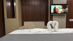 Una cama bien hecha con edredón blanco y manta gris, una toalla blanca dispuesta en forma de corazón, un televisor montado en la pared y un revestimiento de pared de madera.