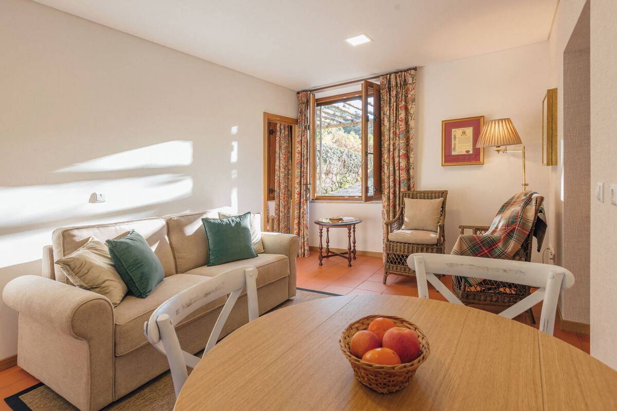 A cozy living room with a sofa, a dining table with a fruit basket, a wicker chair, and a window with curtains.