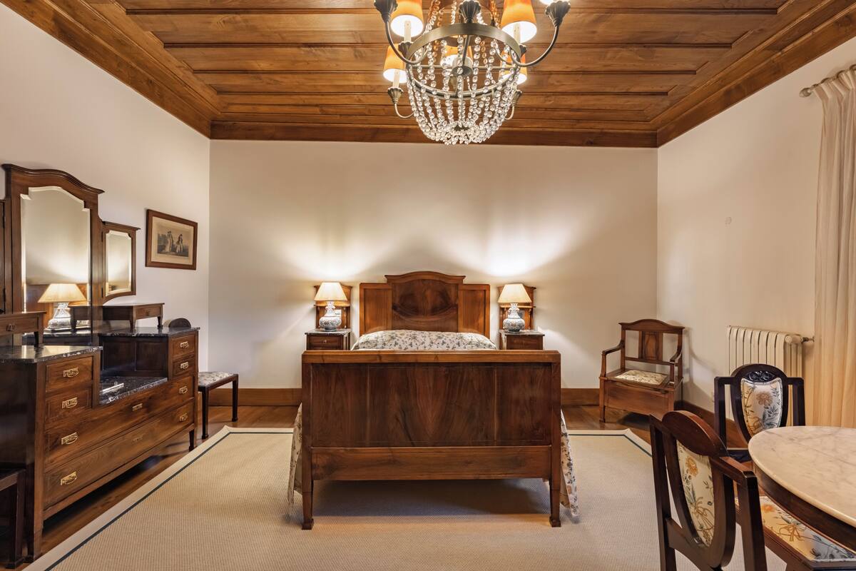 A bedroom with a wooden bed, two bedside tables with lamps, a dresser, a chair, and a chandelier.