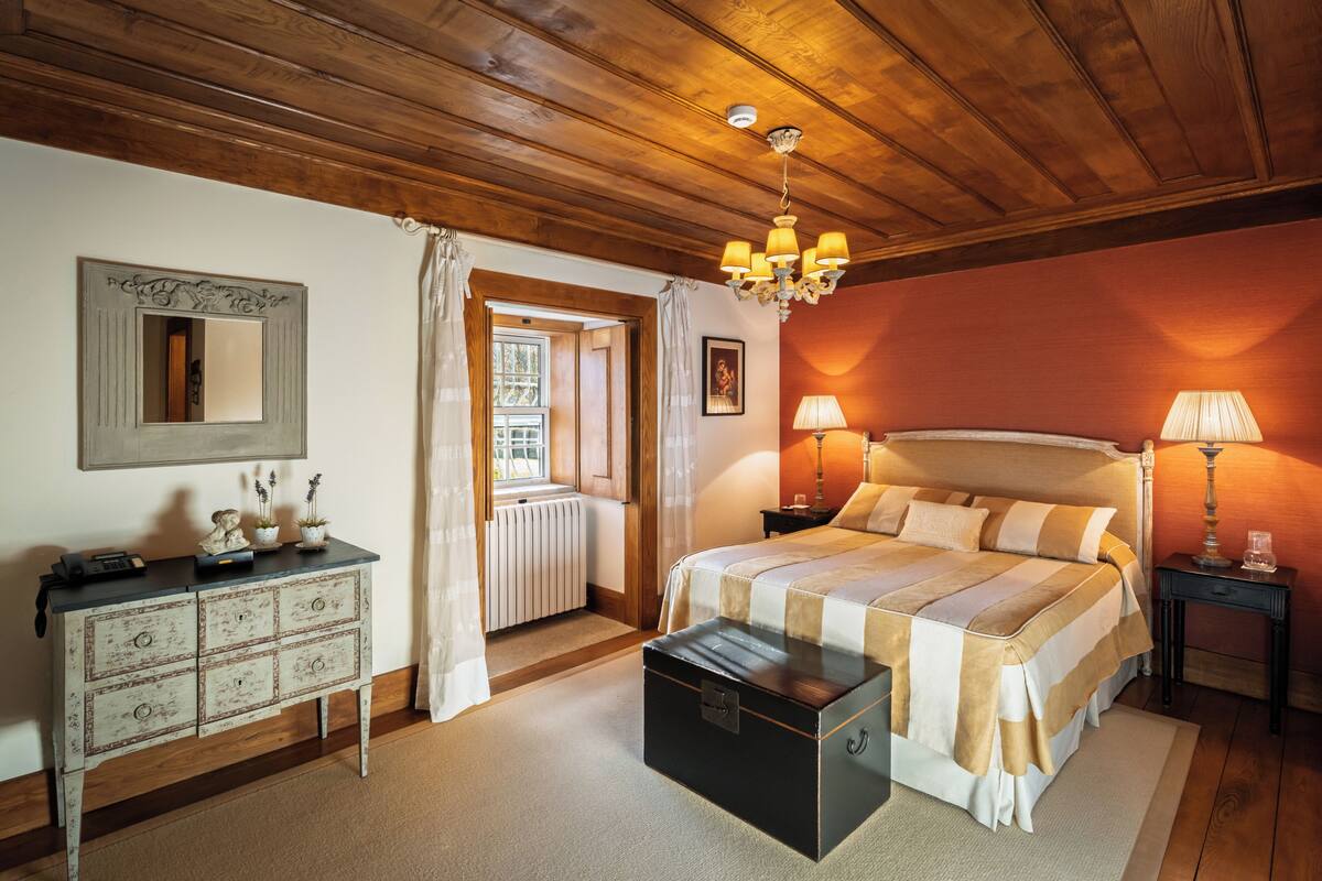 A bedroom with a wooden ceiling, a bed with a striped bedspread, a dresser with drawers, a chest of drawers, and a window with curtains.