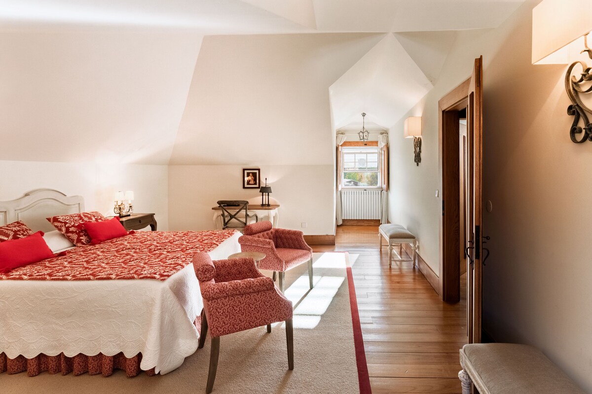 A bedroom with a large bed, two chairs, a small table, and a window with curtains.