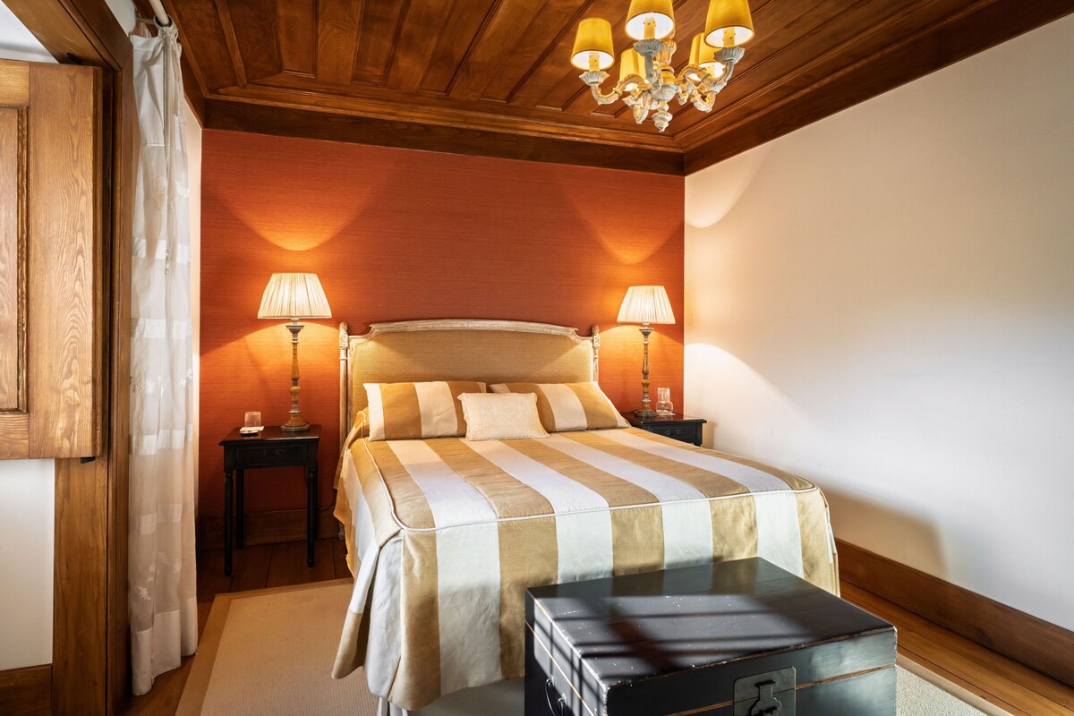 A hotel room with a large bed, two bedside lamps, a wooden headboard, a nightstand, and a trunk.