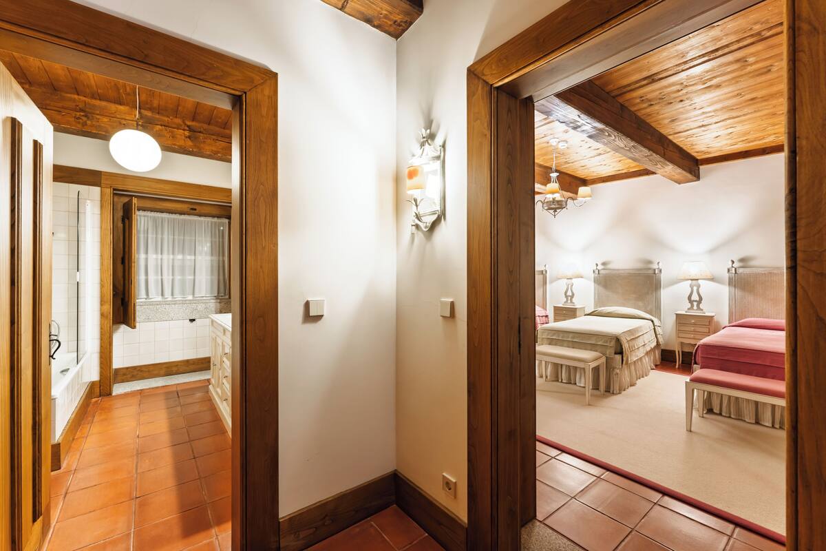 A hallway leading to a bedroom with a wooden ceiling, a bathroom with a shower, and a bed with a pink blanket.