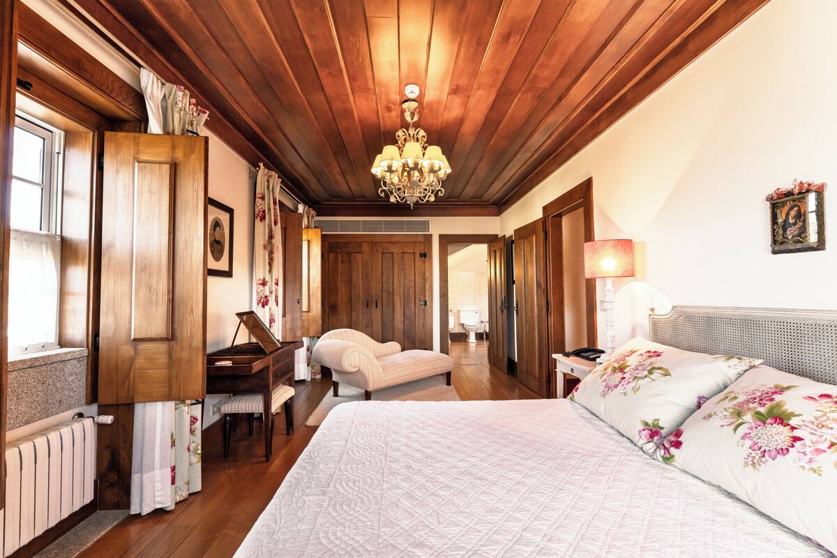 A bedroom with a large bed, wooden ceiling, a piano, a chair, and a desk.
